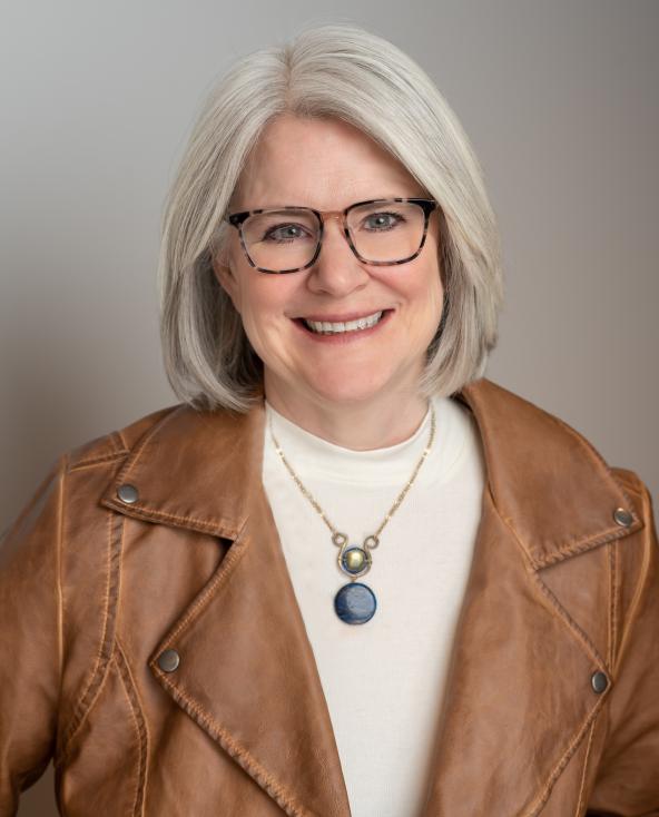 Patti's headshot. Wearing a brown leather jacket and glasses